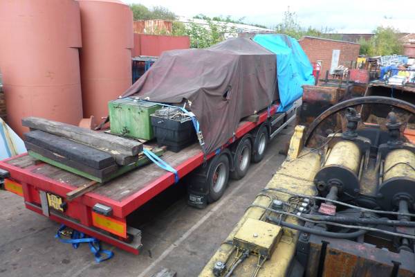 A red flatbed trailer carrying heavy equipment covered by a grey tarpaulin.
