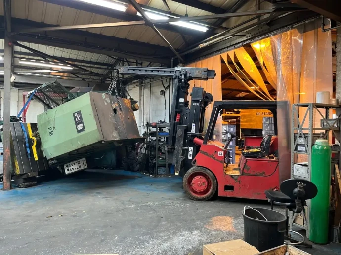 A red forklift lifting a large green industrial press inside a busy workshop.
